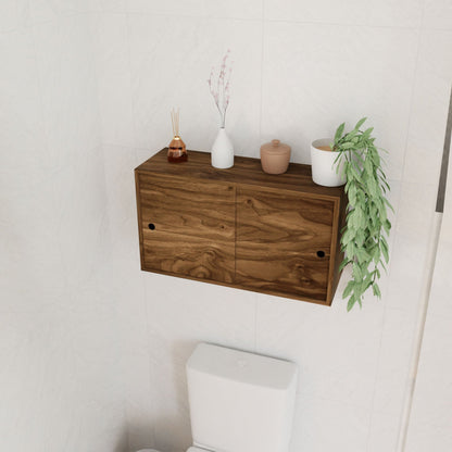 A Krovel Cupboard with Shelf in Walnut, featuring two hand-finished wooden doors, is mounted above a white toilet. The solid cabinet holds decorative items like small vases, a jar, and a lush green plant cascading over the edge. The wall is tiled in white.