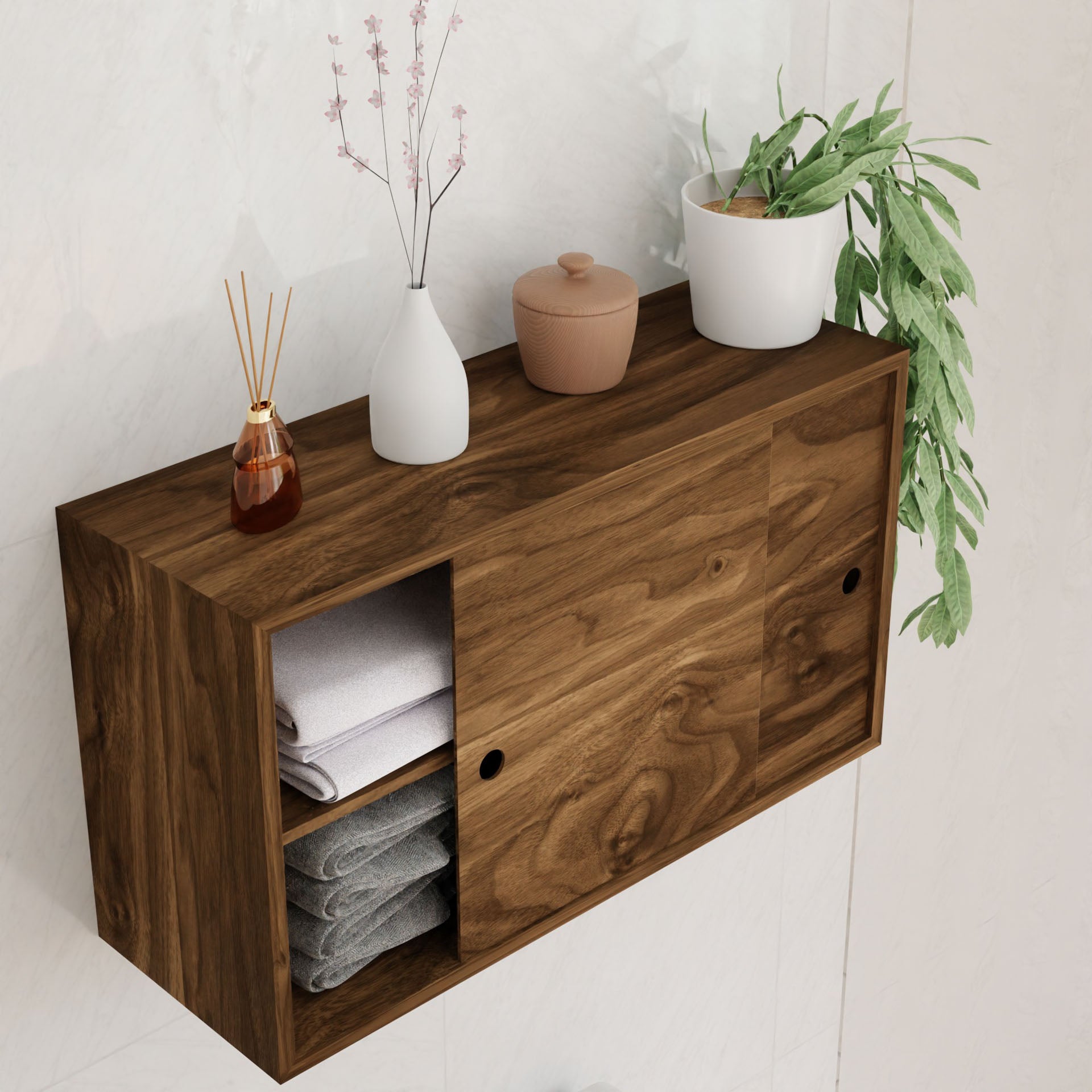 The Krovel "Cupboard with Shelf in Walnut" is a wall-mounted, solid walnut unit that doubles as a floating storage cabinet. Featuring sliding doors, it displays items like a white vase with branches, a reed diffuser, and plants on top while neatly folded towels are visible inside.