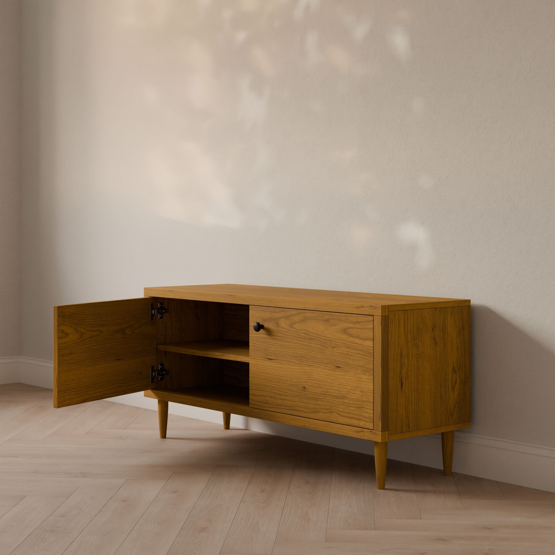 The Double Door Media Cabinet in Cherry by Krovel Furniture Co. stands against a light-colored wall, featuring retro-style splayed legs and crafted from formaldehyde-free hardwood veneer. One door is open, revealing a shelf inside as sunlight casts soft shadows on the wall and floor.