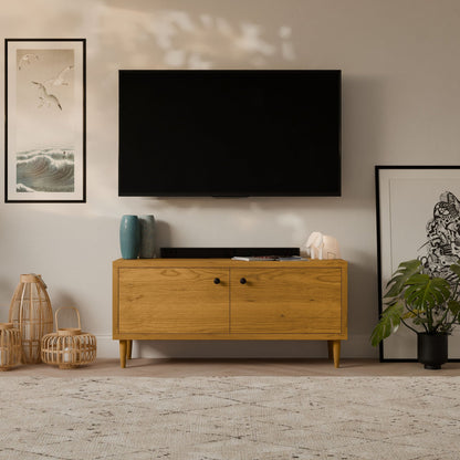 A modern living room showcases a flat-screen TV on a light gray wall, above the Krovel Furniture Co.s Double Door Media Cabinet in Cherry. Two vases, a sculpture, artwork, and plants enhance the decor, while two wicker lanterns rest on the patterned rug to the left.