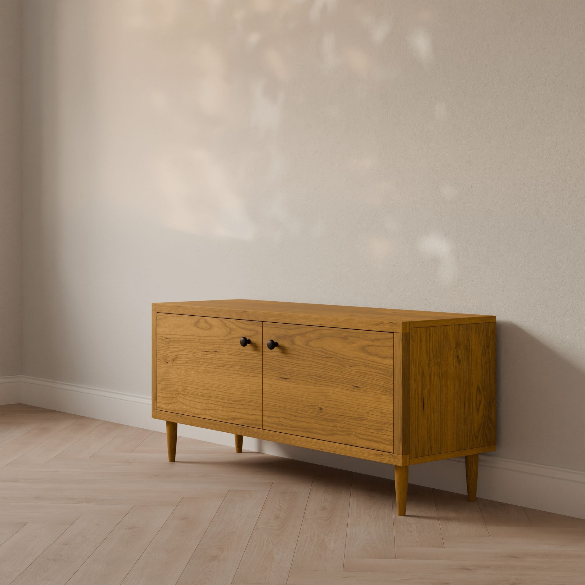 The Double Door Media Cabinet in Cherry by Krovel Furniture Co., crafted from formaldehyde-free hardwood veneer, features two cabinet doors with small black handles. It stands against a light-colored wall, with a light wooden floor enhancing its natural tones as sunlight casts soft shadows.