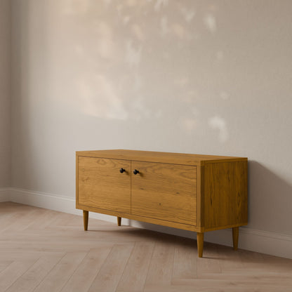 The Double Door Media Cabinet in Cherry by Krovel Furniture Co., crafted from formaldehyde-free hardwood veneer, features two cabinet doors with small black handles. It stands against a light-colored wall, with a light wooden floor enhancing its natural tones as sunlight casts soft shadows.