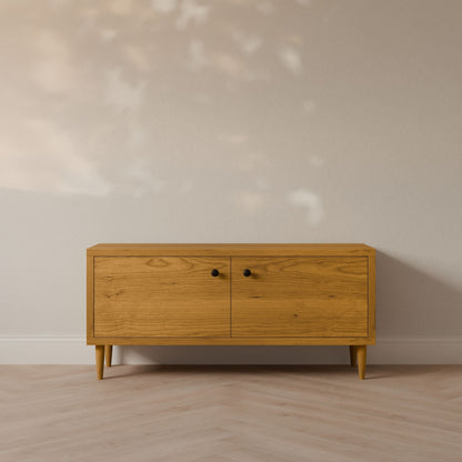 The Double Door Media Cabinet in Cherry, by Krovel Furniture Co., is crafted from premium cabinet grade plywood with formaldehyde-free hardwood veneer. It features two doors with round black handles and angled legs, set against a light wall. The floor has a light wood herringbone pattern.