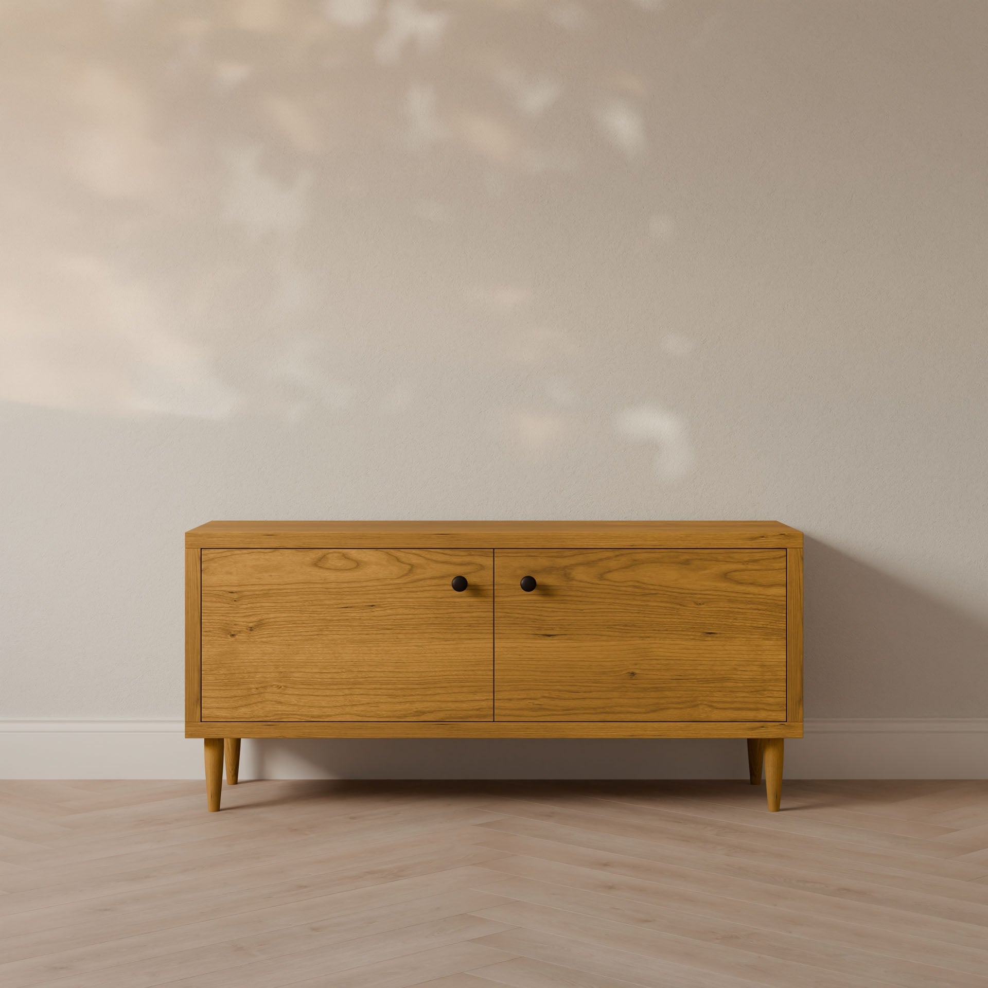 The Maine Made Double Door Media Cabinet by Krovel Furniture Co. stands against a light-colored wall on a chevron-patterned floor, featuring two doors with simple black knobs and finished without formaldehyde. Sunlight dapples the wall behind it.