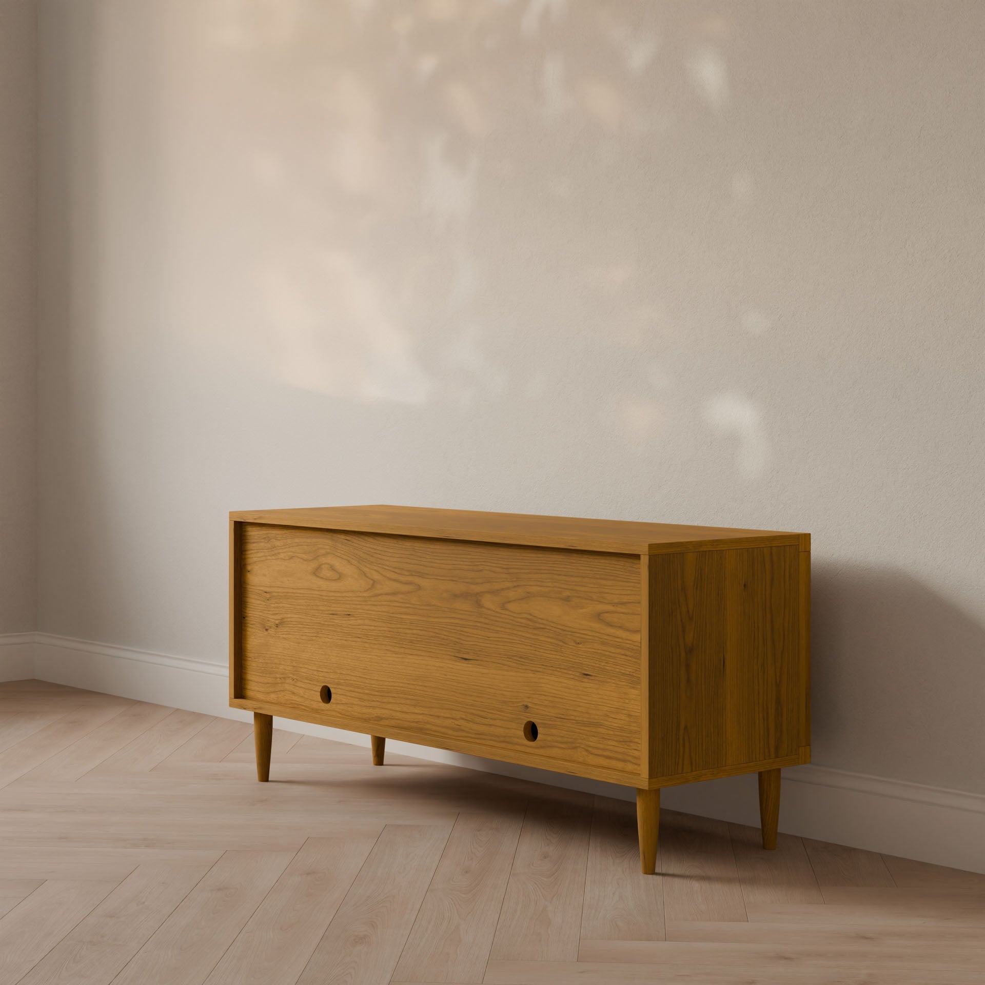 A customizable, formaldehyde-free wooden storage bench with angled legs sits against a light wall on wood flooring. Warm sunlight sets a minimalist mood—perfect with the Maine Made Double Door Media Cabinet by Krovel Furniture Co.