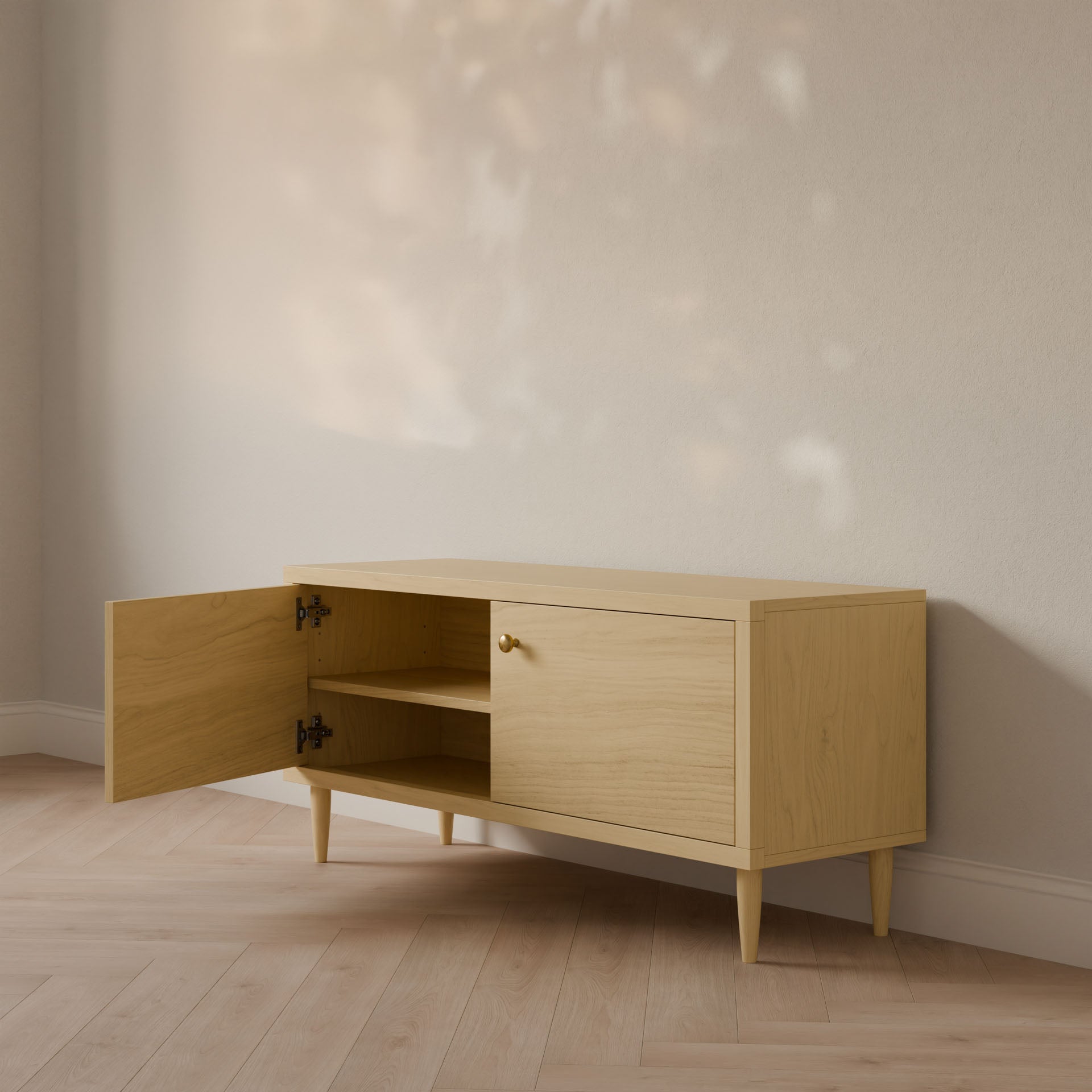 The Double Door Media Cabinet in Maple by Krovel Furniture Co. is a minimalist, mid-century modern piece handmade in Portland, Maine. Crafted from formaldehyde-free hardwood with two doors—one ajar to show its shelves—its positioned against a light wall with sunlight casting leaf-patterned shadows.