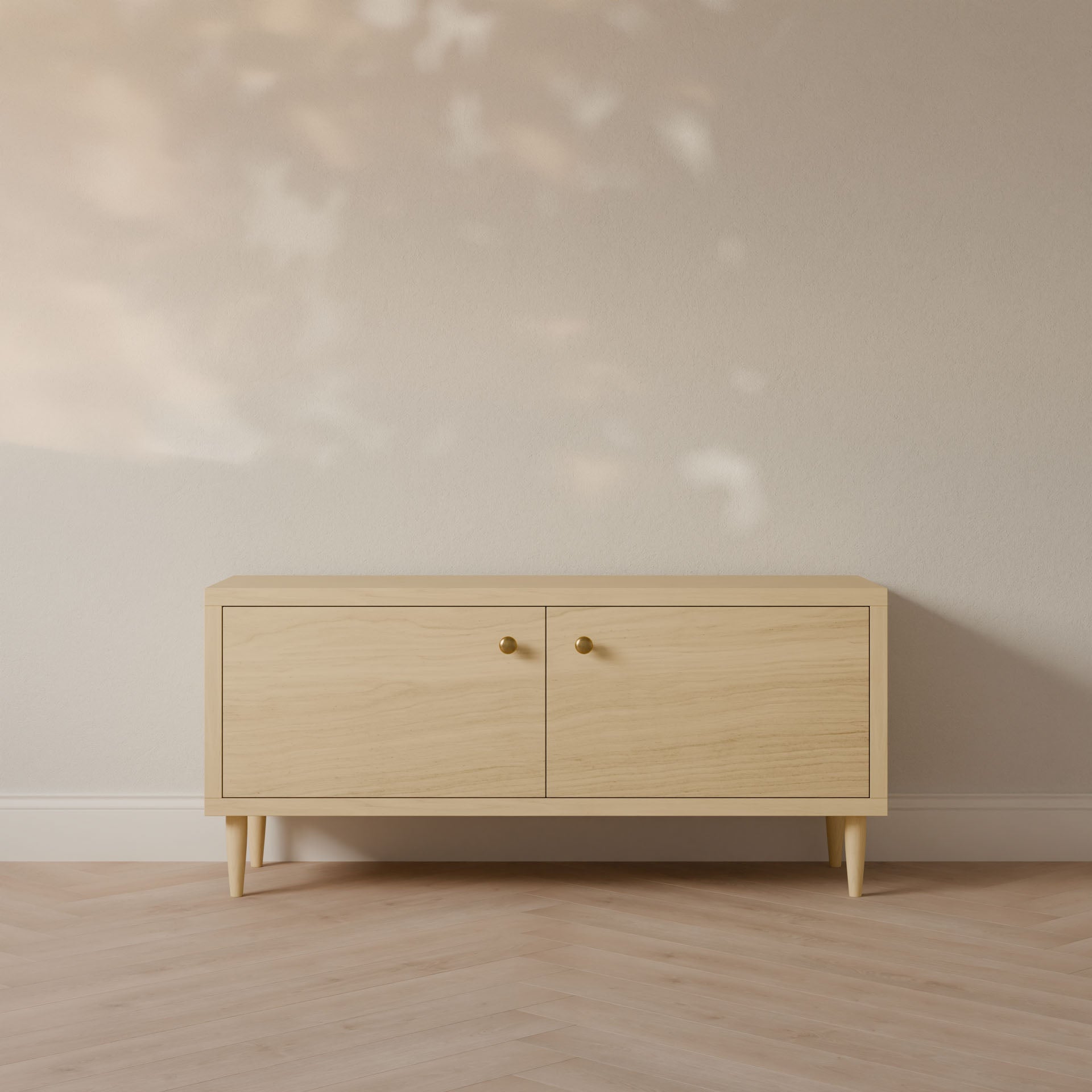 The Double Door Media Cabinet in Maple by Krovel Furniture Co. is a handmade sideboard from Portland, Maine, featuring a minimalist design with a light natural finish. Crafted from formaldehyde-free hardwood, it showcases round brass knobs and stands elegantly on tapered legs against a beige wall and herringbone floor.