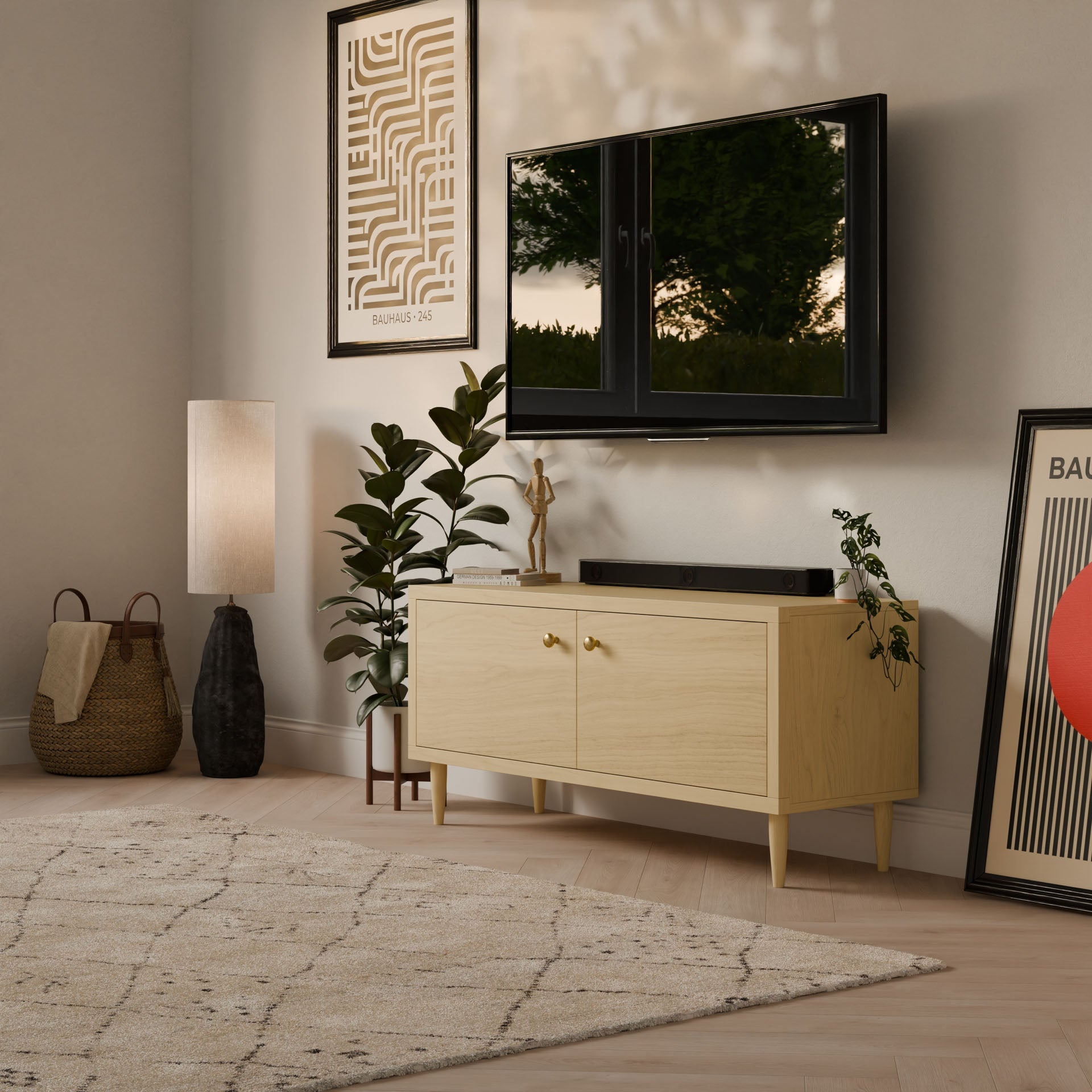 A cozy living room features a wall-mounted TV above a Double Door Media Cabinet in Maple by Krovel Furniture Co., handmade in Portland, Maine. Decor adorns the cabinet, with a potted plant and lamp adding warmth. Abstract art leans against the wall, along with a woven basket and patterned rug.