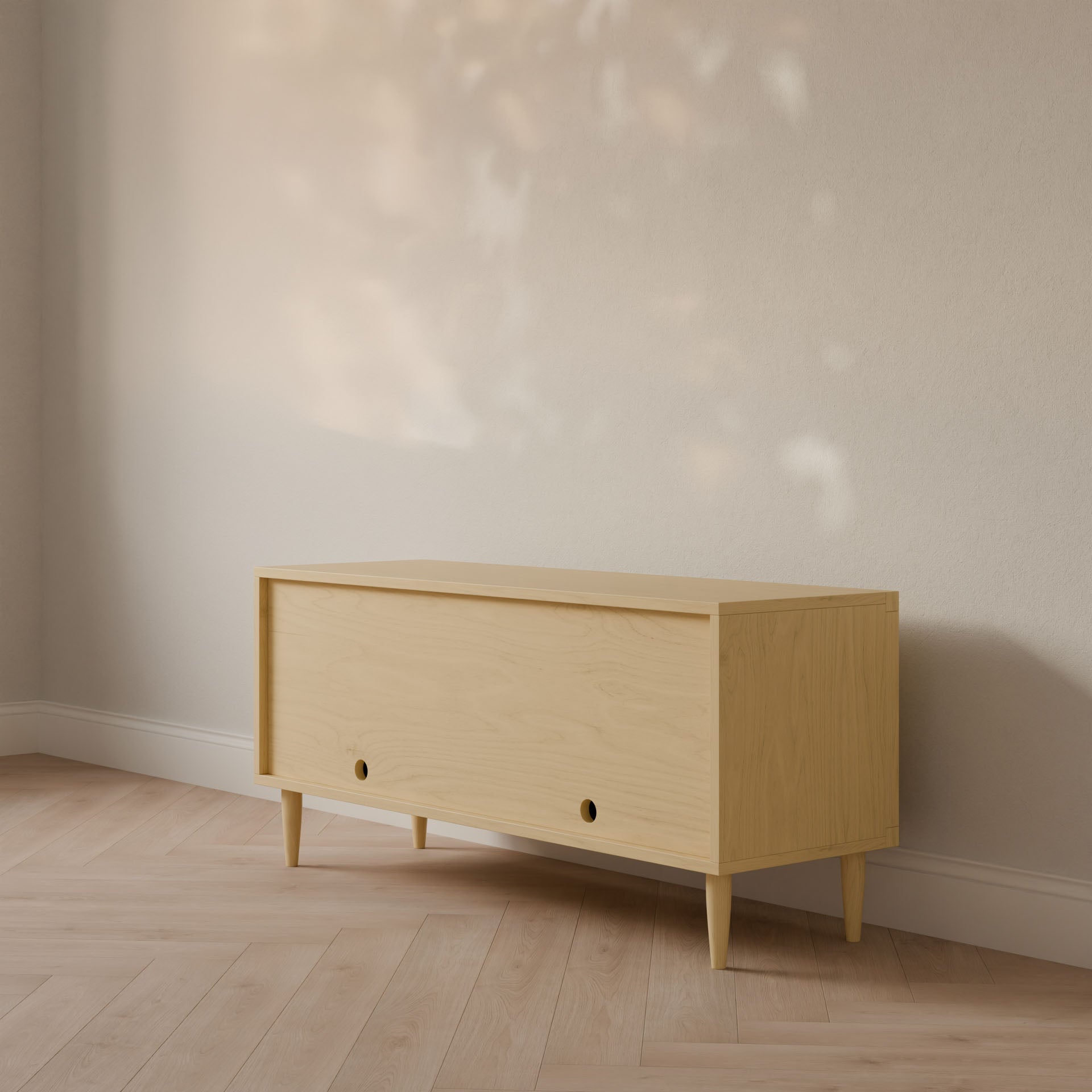 A Double Door Media Cabinet made of formaldehyde-free maple wood, handcrafted by Krovel Furniture Co. in Portland, Maine, stands against a beige wall with sunlight filtering through the window, casting shadows on the wooden floor. It features a minimalist design with two circular front openings.
