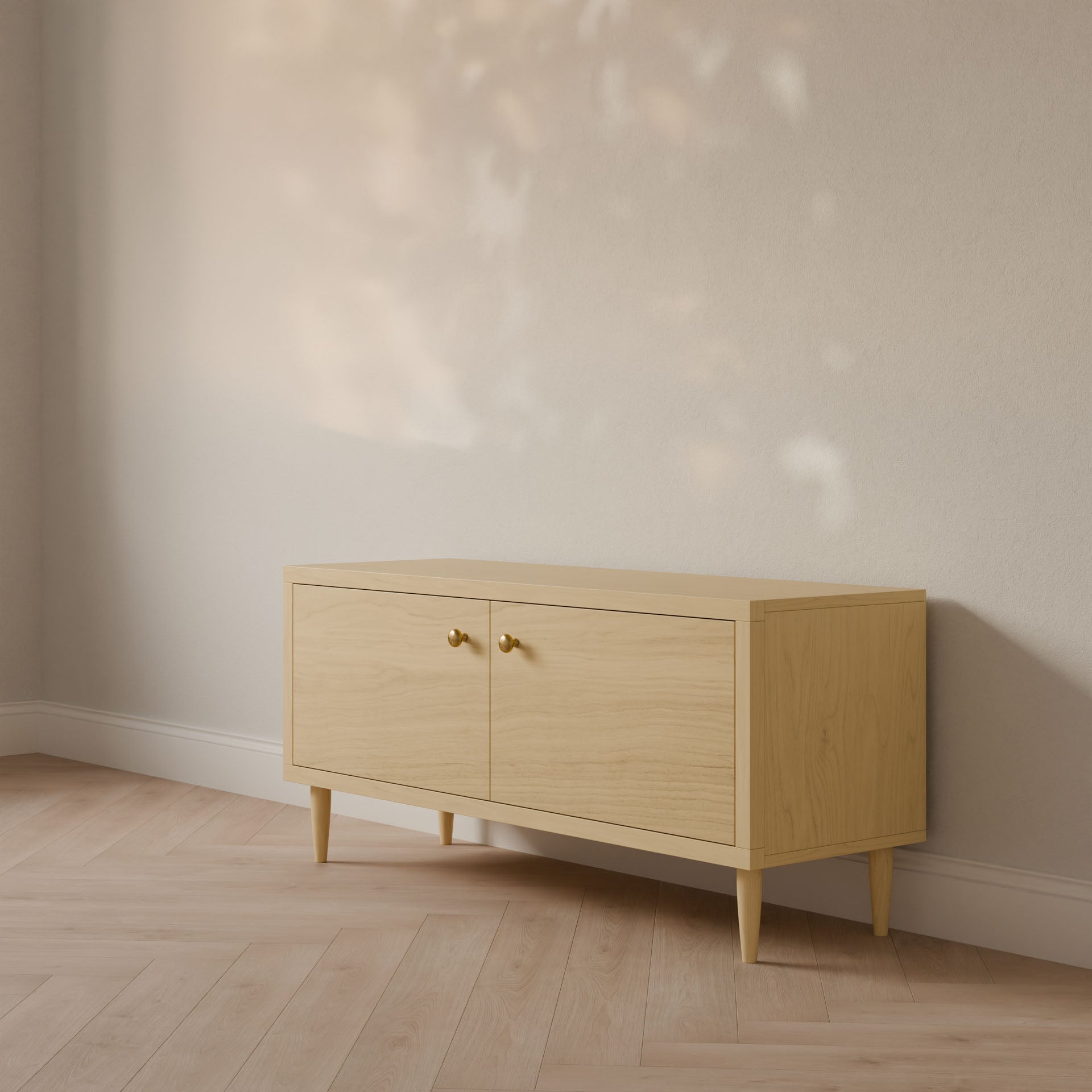 A Double Door Media Cabinet in Maple by Krovel Furniture Co. rests against a cream wall, featuring two doors with gold handles. Sunlight creates soft shadows on the formaldehyde-free hardwood floor that matches the cabinets hue. Crafted in Portland, Maine.