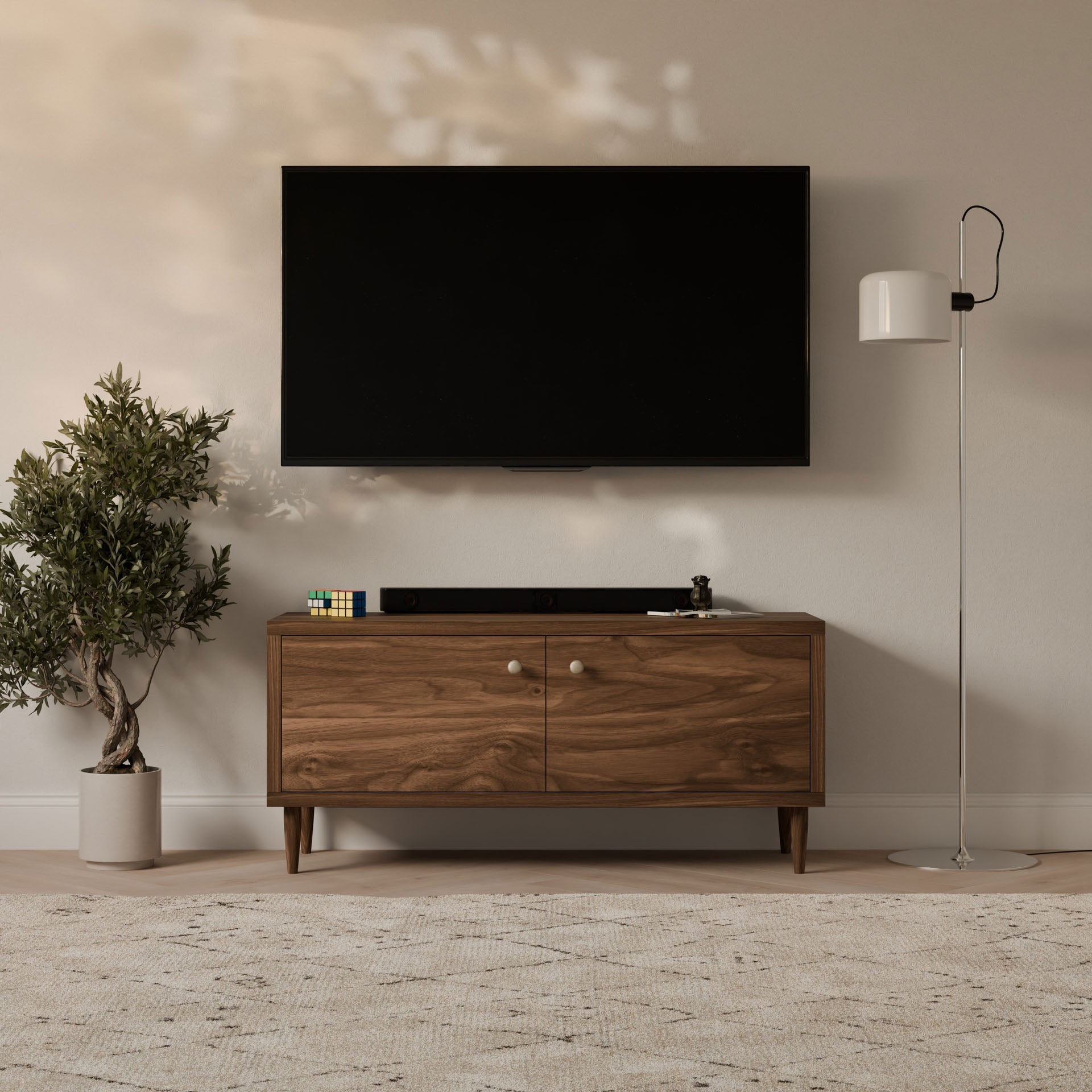 A modern living room setup includes a wall-mounted TV above a handmade Double Door Media Cabinet in Walnut by Krovel Furniture Co. A potted plant is on the left, a floor lamp on the right, and decorative items adorn the console. A light-colored rug covers the formaldehyde-free hardwood floor.