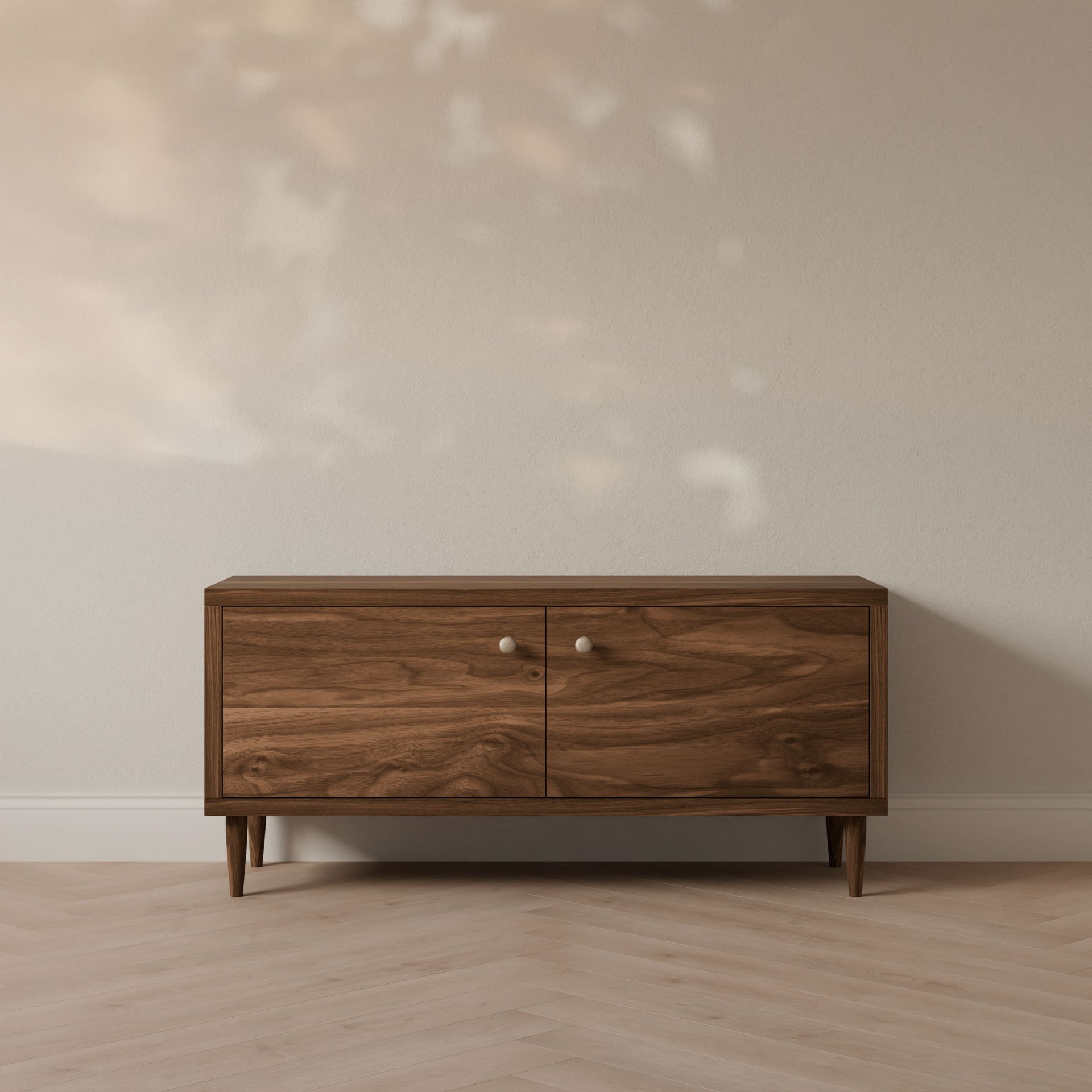 The Double Door Media Cabinet in Walnut by Krovel Furniture Co., a handmade mid-century modern piece from Portland, Maine, features formaldehyde-free hardwood with two doors and round knobs. It stands against a neutral wall with dancing leafy shadows on a herringbone-patterned floor.