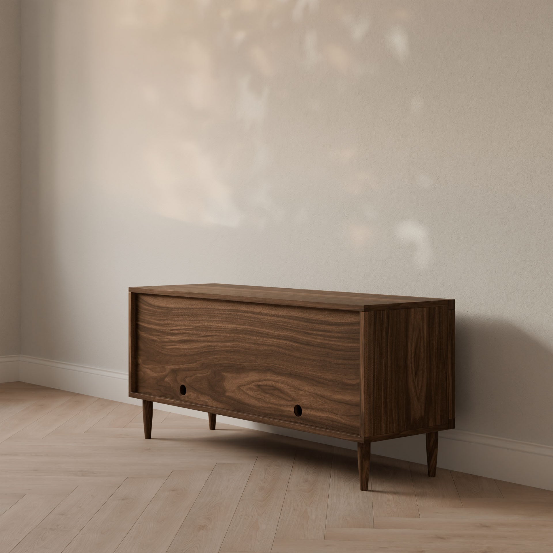 A Double Door Media Cabinet in Walnut by Krovel Furniture Co. stands against a neutral wall, featuring tapered legs and circular handles. Sunlight through a window casts dappled shadows on the formaldehyde-free hardwood floor, highlighting its mid-century modern design.