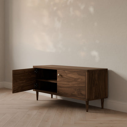 Against a beige wall, the elegant Double Door Media Cabinet in Walnut by Krovel Furniture Co. showcases its handmade design from Portland, Maine. Soft shadows from a window highlight its formaldehyde-free hardwood finish, complementing the light-colored wooden floor and open-door shelving.