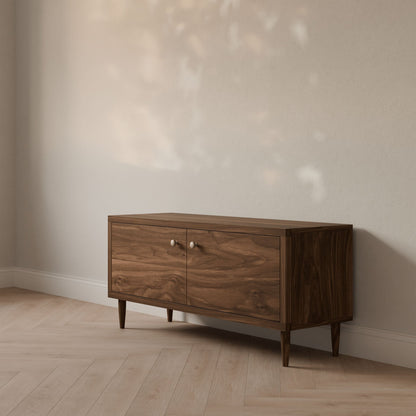 The Double Door Media Cabinet in Walnut by Krovel Furniture Co., handmade in Portland, Maine, features sleek legs and two doors. It stands against a light wall on pale wood flooring, with sunlight filtering through to create dappled shadows.