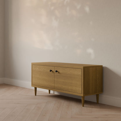 A handmade Double Door Media Cabinet in White Oak by Krovel Furniture Co. stands against a light-colored wall in Portland, Maine. The rooms light wood floor and dappled sunlight create patterns on this customizable sideboard with two closed cabinets.