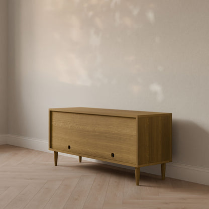 The Double Door Media Cabinet in White Oak by Krovel Furniture Co., handmade in Portland, Maine, stands against a neutral wall on a light wood floor. Soft, dappled sunlight patterns above add warmth and texture to the minimalistic setting.