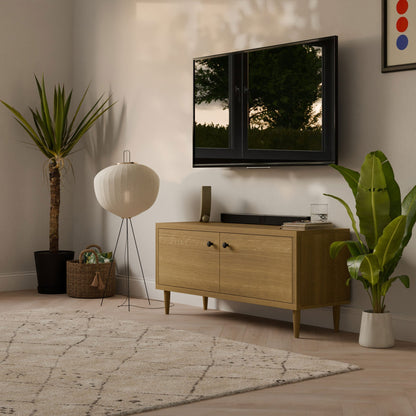 A modern living room includes a Krovel Furniture Co. Double Door Media Cabinet in White Oak with a wall-mounted TV above. Nearby, a tall plant, wicker basket, tall lamp, and vibrant wall art enhance this stylish space. The cabinet is crafted with formaldehyde-free hardwood veneer.