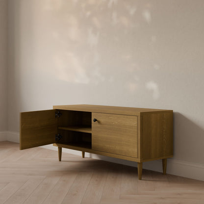 The Maine Made Double Door Media Cabinet by Krovel Furniture Co. stands against a light wall, one door open to show shelves inside. Sunlight casts patterns on the wall and a light wood floor—ideal for those who love customizable media cabinets.