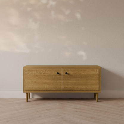The Maine Made Double Door Media Cabinet by Krovel Furniture Co. features two doors with black knobs, is formaldehyde-free, and stands against a light beige wall on a herringbone-patterned floor—ideal for customizable media setups.
