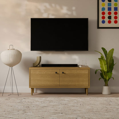 A modern living room features a wall-mounted flat-screen TV above a customizable Double Door Media Cabinet in White Oak, handmade by Krovel Furniture Co. in Portland, Maine. Decor includes glass art, books, and sculptures with a floor lamp and plant on each side and colorful abstract artwork for flair.