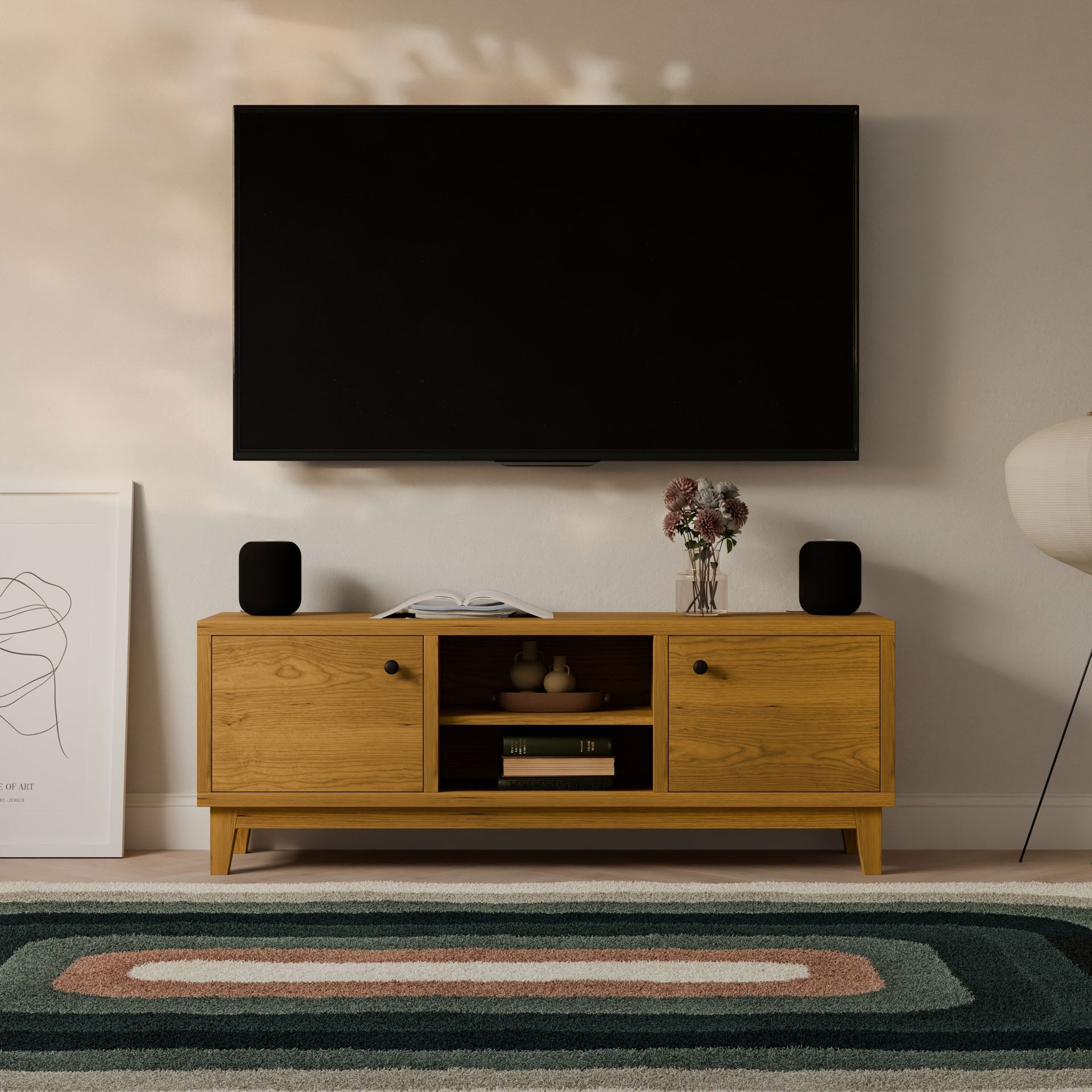 A flat-screen TV is mounted above a Krovel Furniture Co. Maine Made Double Door, Open Shelf Media Cabinet with books, vases, and black speakers. A framed abstract art piece, a lamp, and a patterned rug complete the scene.