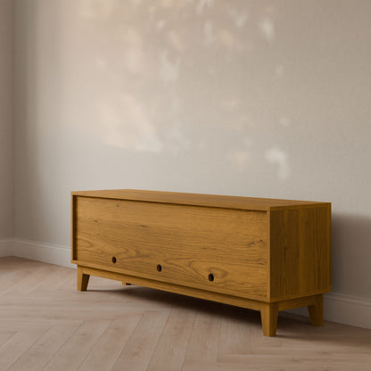 The Maine Made Double Door, Open Shelf Media Cabinet by Krovel Furniture Co. features a formaldehyde-free wooden design with closed front doors and three round knobs, positioned against a light wall on a herringbone floor in natural light.