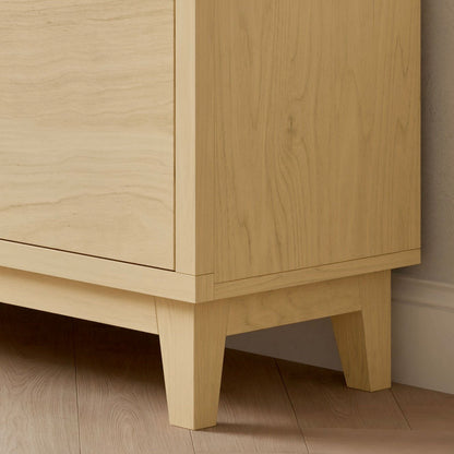 Close-up of the lower corner of the Maine Made Double Door, Open Shelf Media Cabinet by Krovel Furniture Co., crafted from light wood with straight legs, placed on a light wood floor beside a white wall.