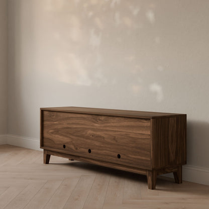 The Double Door, Open Shelf Media Cabinet in Walnut by Krovel Furniture Co. stands on a light wooden floor against a beige wall. Sunlight from a nearby window casts soft shadows onto the wall, enhancing the warmth and cozy atmosphere with its minimalist design crafted from hardwood veneer.