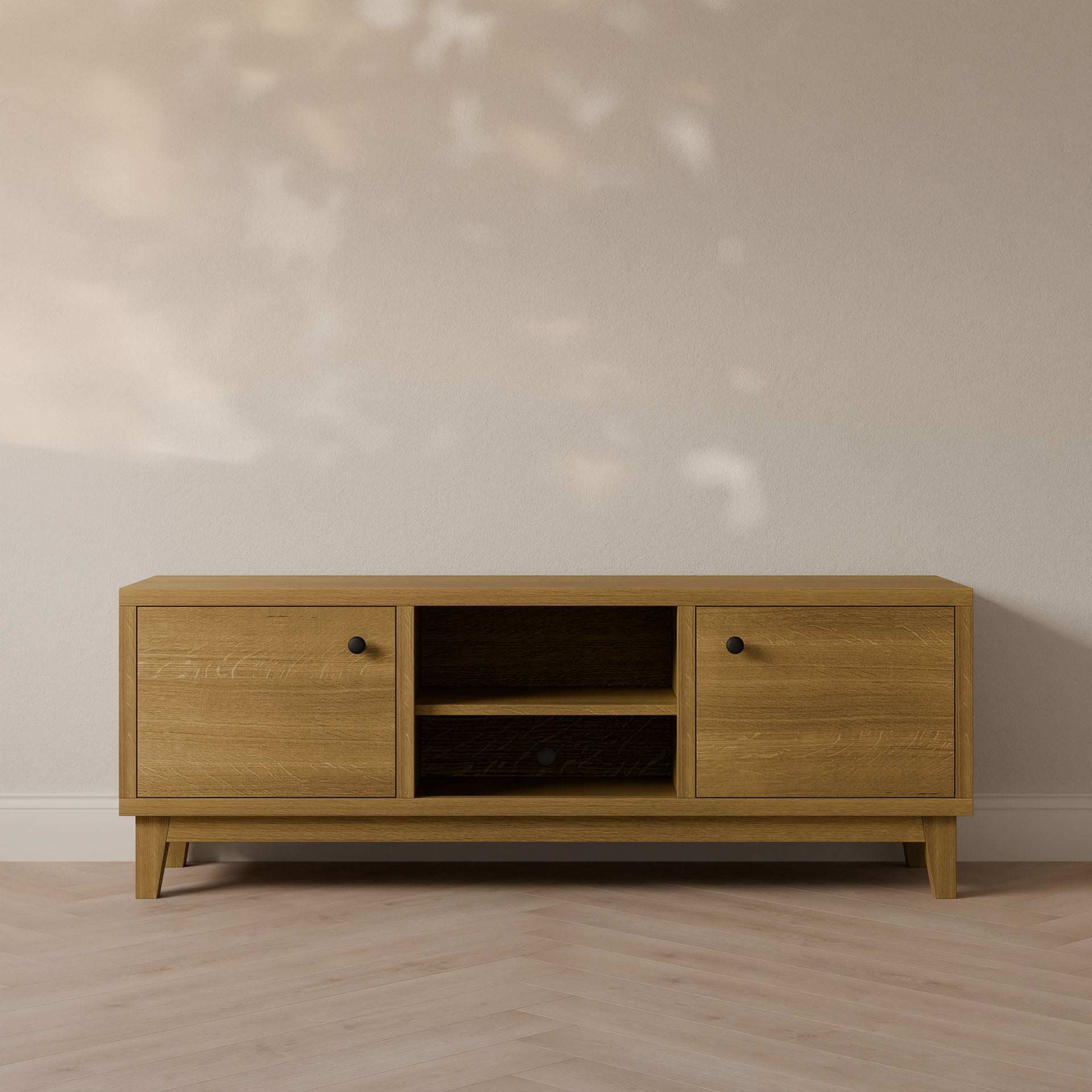 The Double Door, Open Shelf Media Cabinet in White Oak by Krovel Furniture Co. rests against a light wall, showcasing two cabinets with black knobs and central open shelving. Its minimalist design pairs beautifully with the floors subtle herringbone pattern and soft light casting gentle shadows.