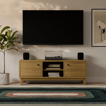A modern living room features a wall-mounted TV above the Maine Made Double Door, Open Shelf Media Cabinet from Krovel Furniture Co., flanked by two black speakers. A plant, framed artwork, and a green patterned rug add finishing touches.