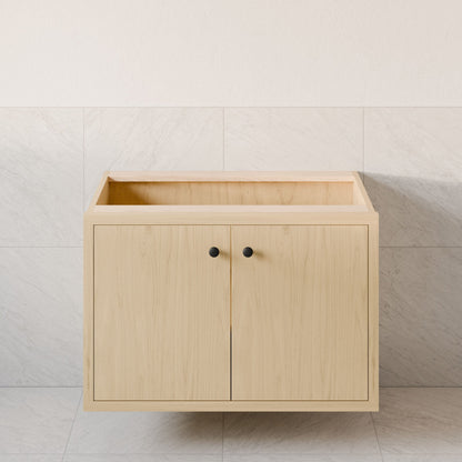 The Double Door Vanity in Maple by Krovel Furniture Co. is a handmade, light wood floating vanity with two black round-handled doors, shown against large light tiles and pictured without a countertop or sink.