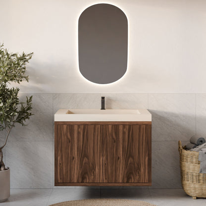 The Maine Made Double Door Vanity by Krovel Furniture Co. features a wood cabinet and cream countertop with a minimalist faucet. An oval backlit mirror hangs above, while a wicker basket with towels sits beside a leafy plant on tiled floors.