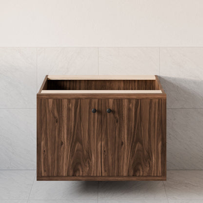 The Maine Made Double Door Vanity by Krovel Furniture Co., a floating hardwood bathroom cabinet with black knobs and two doors, is mounted against a tiled wall without its countertop or sink.