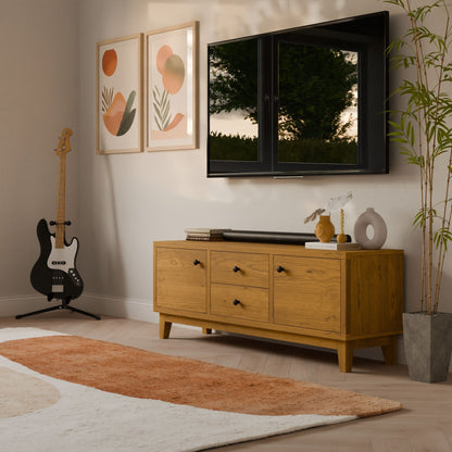 A modern living room showcases a Krovel Furniture Co. Double Drawer, Double Door Media Cabinet in Cherry with soft-close slides. It features a potted plant, a wall-mounted TV, abstract art above, and a guitar in the corner. A brown and white rug accents the light floor while sunlight highlights the hardwood veneer finish.