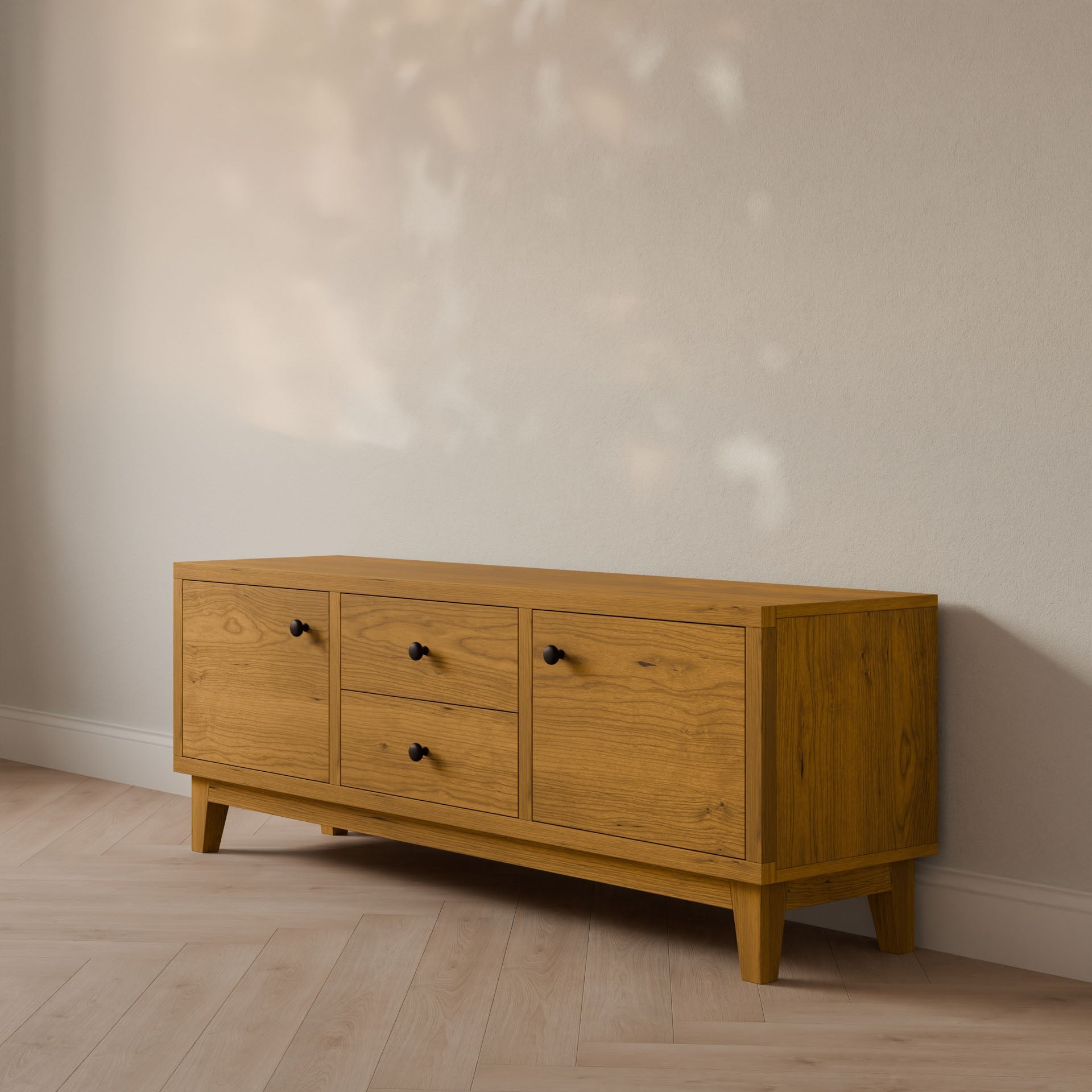 The Double Drawer, Double Door Media Cabinet in Cherry from Krovel Furniture Co. showcases a smooth hardwood veneer with natural wood grain against a light wall. Soft natural light highlights its elegant design, complete with soft-close slides for quiet functionality.