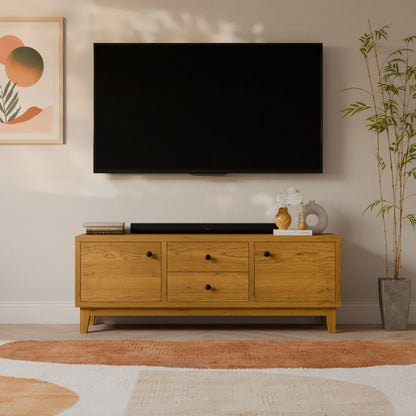 A modern living room setup features a large flat-screen TV mounted on a white wall above the Krovel Furniture Co.s Double Drawer, Double Door Media Cabinet in Cherry. Minimalist artwork and a potted plant enhance the décor, complemented by a textured orange and white rug on the floor.