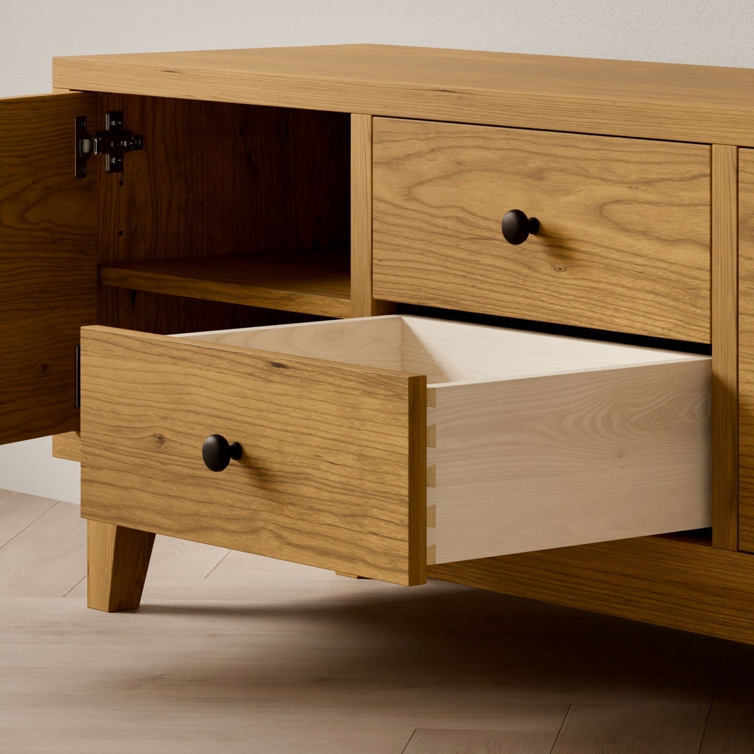 The Double Drawer, Double Door Media Cabinet in Cherry by Krovel Furniture Co. features two open dovetail-jointed drawers, a shelved section, and black round handles. Made from hardwood veneer, its set against a light wall on a hardwood floor.