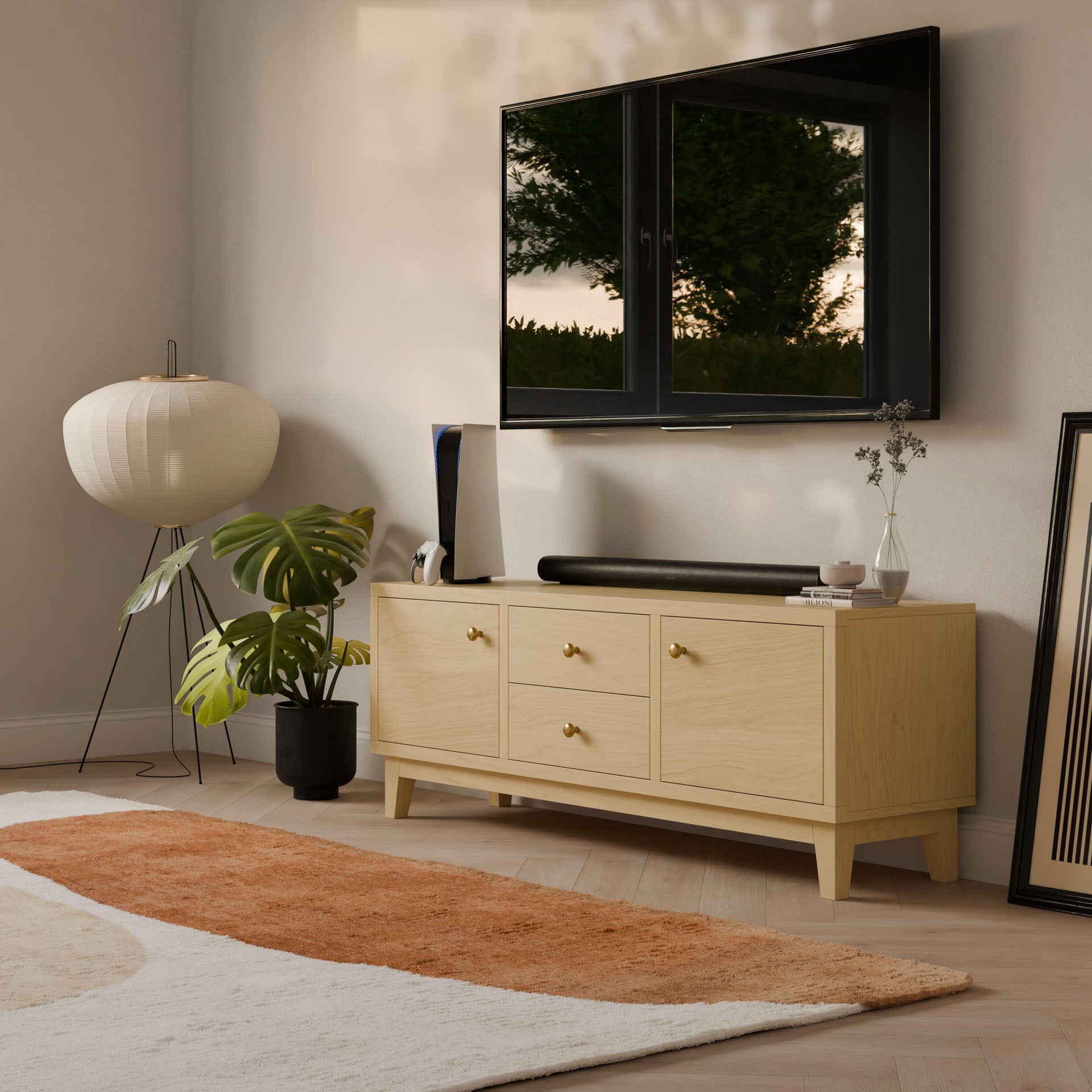 A modern living room features a wall-mounted TV above a Krovel Furniture Co.s Double Drawer, Double Door Media Cabinet in Maple, adorned with gold handles. A leafy plant enhances the space beside a floor lamp, while a white and brown abstract rug covers the wooden floor, and a framed picture leans on the wall.