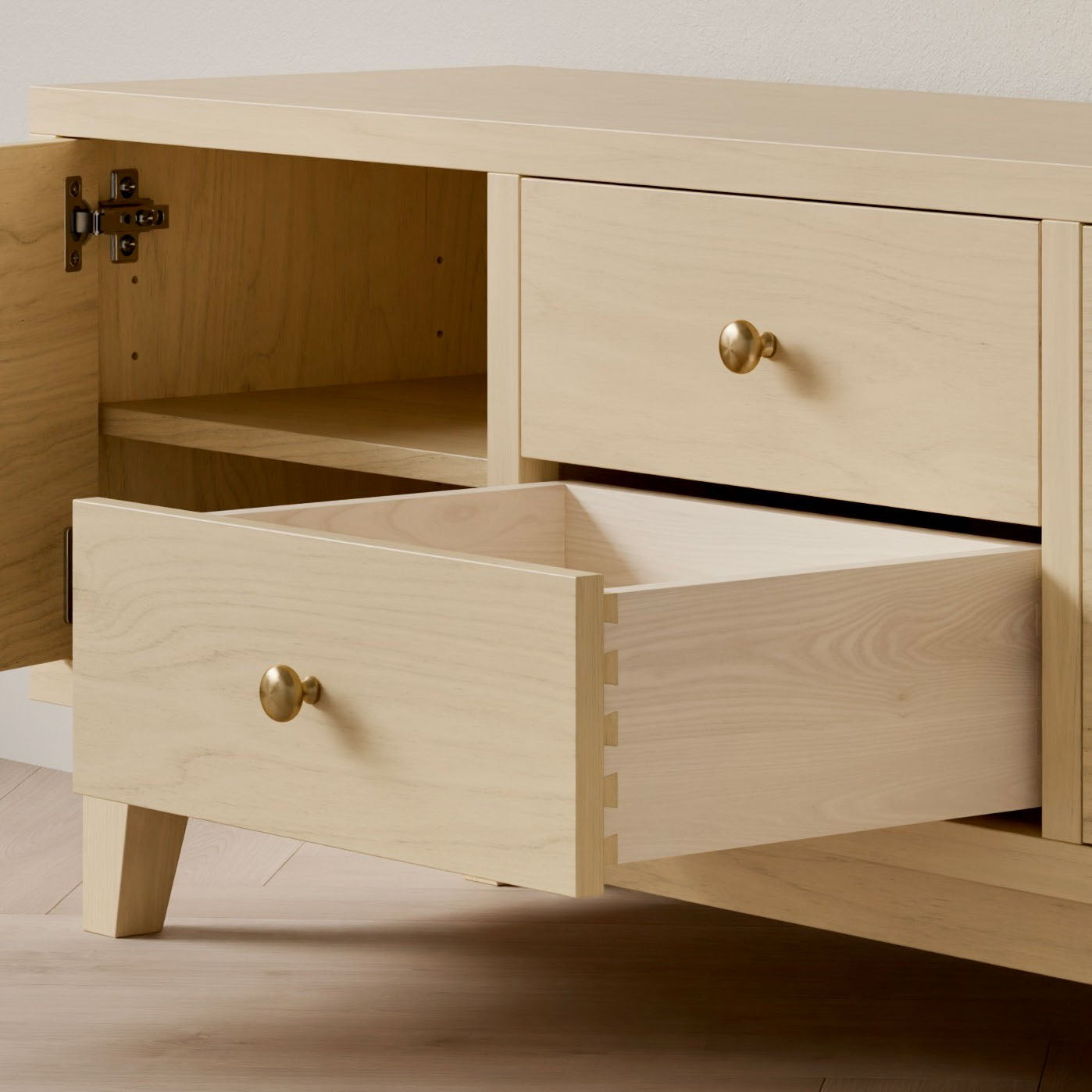 The Maine Made Double Drawer, Double Door Media Cabinet by Krovel Furniture Co. is a customizable light wood sideboard with round brass knobs, handmade in Portland, ME. It features angled legs and open compartments for storage.