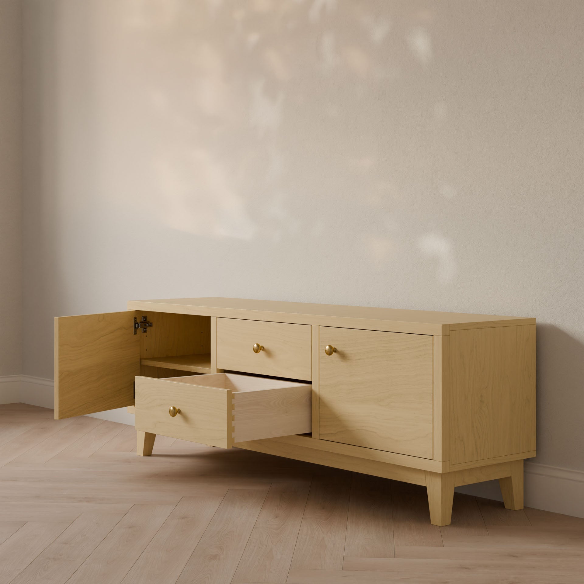 The Double Drawer, Double Door Media Cabinet in Maple by Krovel Furniture Co. is a light wooden sideboard with two cabinets and an open drawer on the left. It features small brass handles, angled legs, and stands against a cream wall on a light hardwood floor, perfectly embodying customizable media cabinet charm.