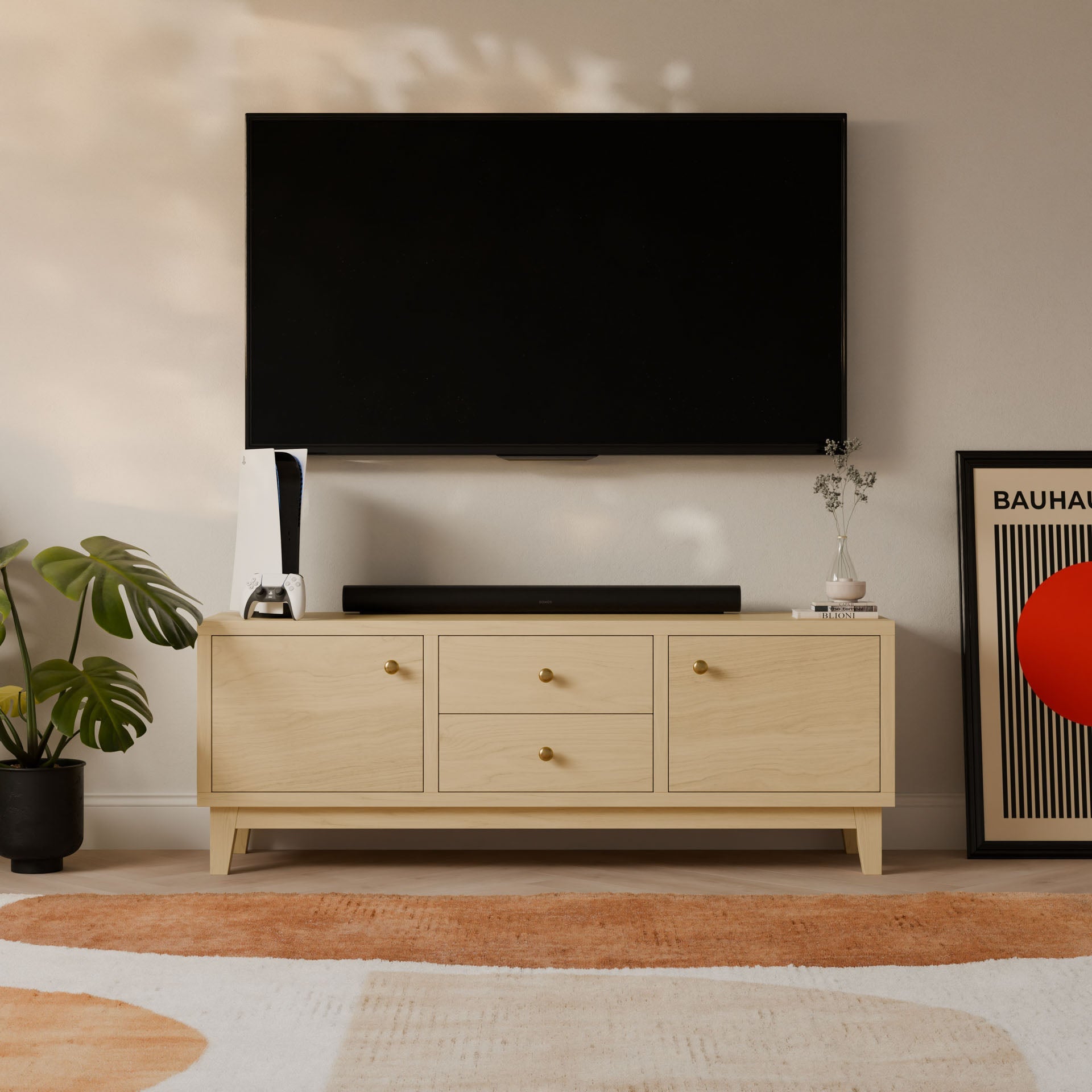 A modern living room showcases a wall-mounted TV above a Krovel Furniture Co. Double Drawer, Double Door Media Cabinet in Maple, handmade in Portland, Maine. The cabinet holds a speaker and decor. A potted plant and minimalist geometric art flank the setup, enhanced by a patterned rug on the floor.