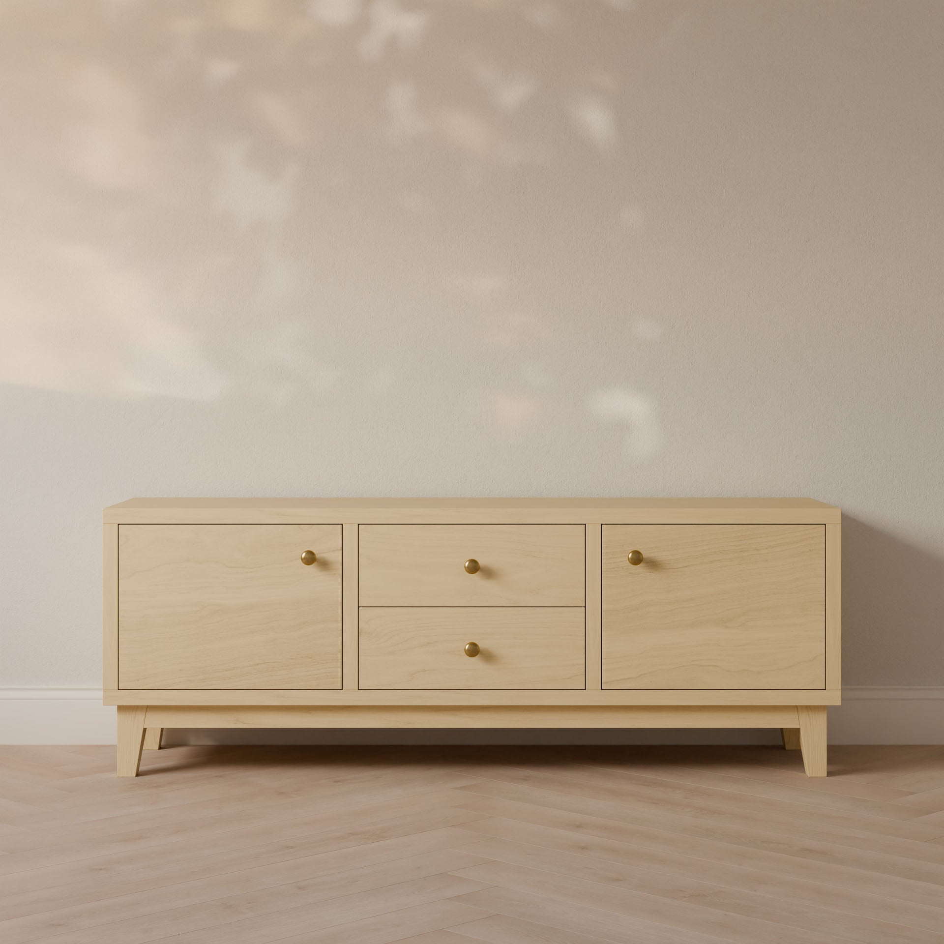 The Double Drawer, Double Door Media Cabinet in Maple by Krovel Furniture Co. is a minimalist piece handmade in Portland, Maine. It features two doors on the left and two drawers on the right with brass knobs. Set on angled splayed legs, this cabinet adds style to any room against a neutral wall with leaf-shadow patterns.