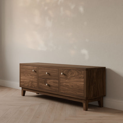 The Double Drawer, Double Door Media Cabinet in Walnut by Krovel Furniture Co. stands on a wooden floor against a white wall, with soft light and leaf-shaped shadows dancing above to highlight its elegant design.
