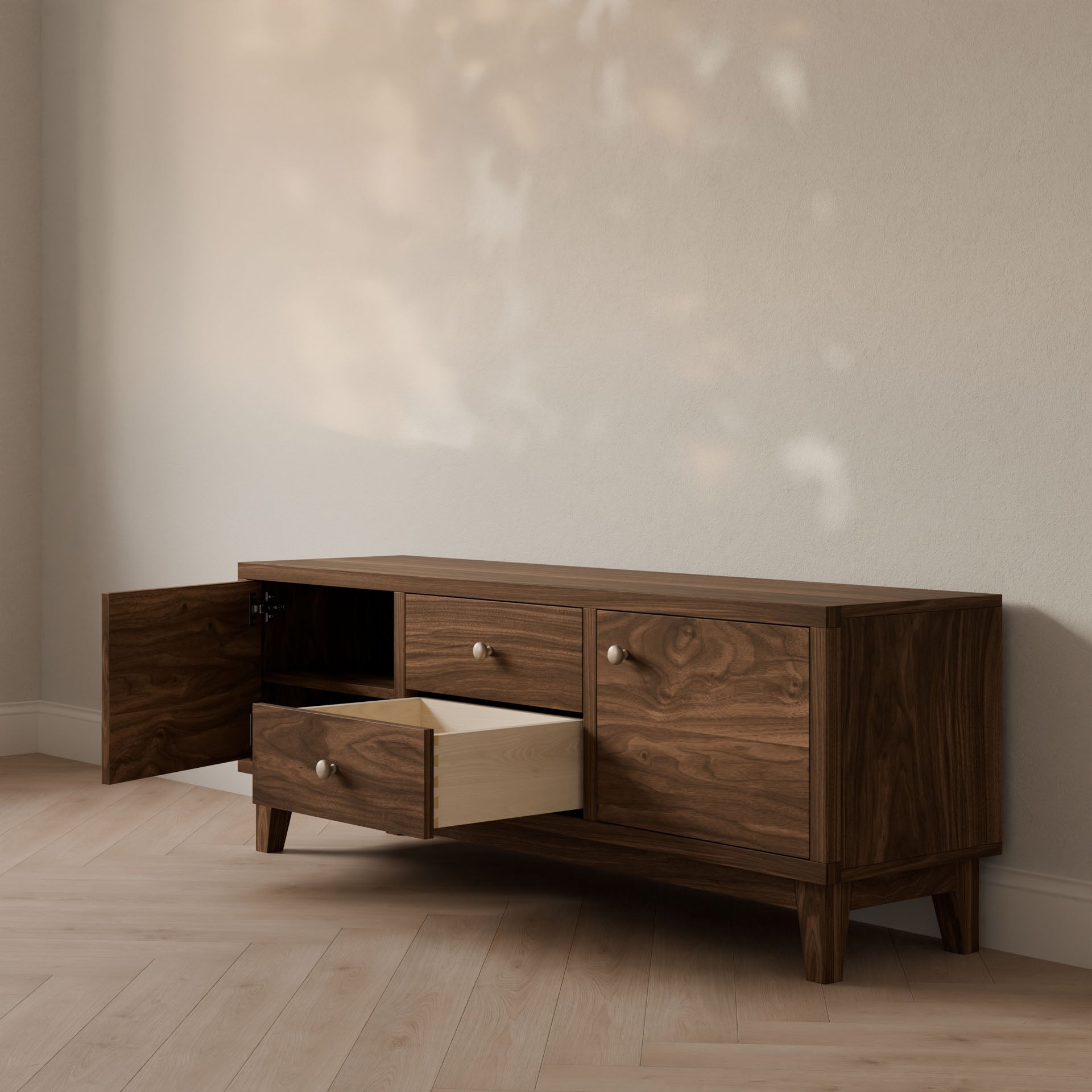 A dark walnut media cabinet by Krovel Furniture Co. sits against a light wall, featuring two solid maple drawers and two doors. One drawer and one door open to show the light wood interior, while tree leaf shadows play on the light wood floor.