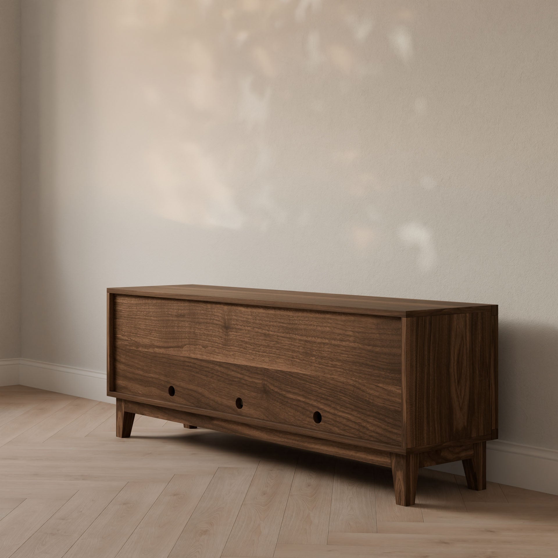 The Double Drawer, Double Door Media Cabinet in Walnut by Krovel Furniture Co. stands against a light-colored wall. It features handmade hardwood veneer, circular cutout handles on the two large drawers, and rests on short legs, with sunlight creating reflections behind it.