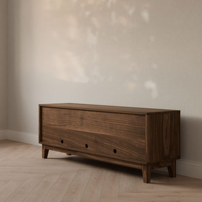 The Maine Made Double Drawer, Double Door Media Cabinet by Krovel Furniture Co. stands against a light wall on a herringbone floor, showcasing clean lines, two circular handles, and customizable features in soft natural light.