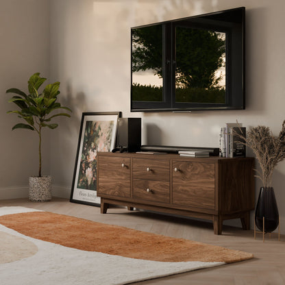 A modern living room showcases a sleek wall-mounted TV above a Krovel Furniture Co. Double Drawer, Double Door Media Cabinet in Walnut. A potted plant and framed artwork are nearby, with a plush rug featuring earthy tones on the light wood floor and decorative items adorning the media cabinet.