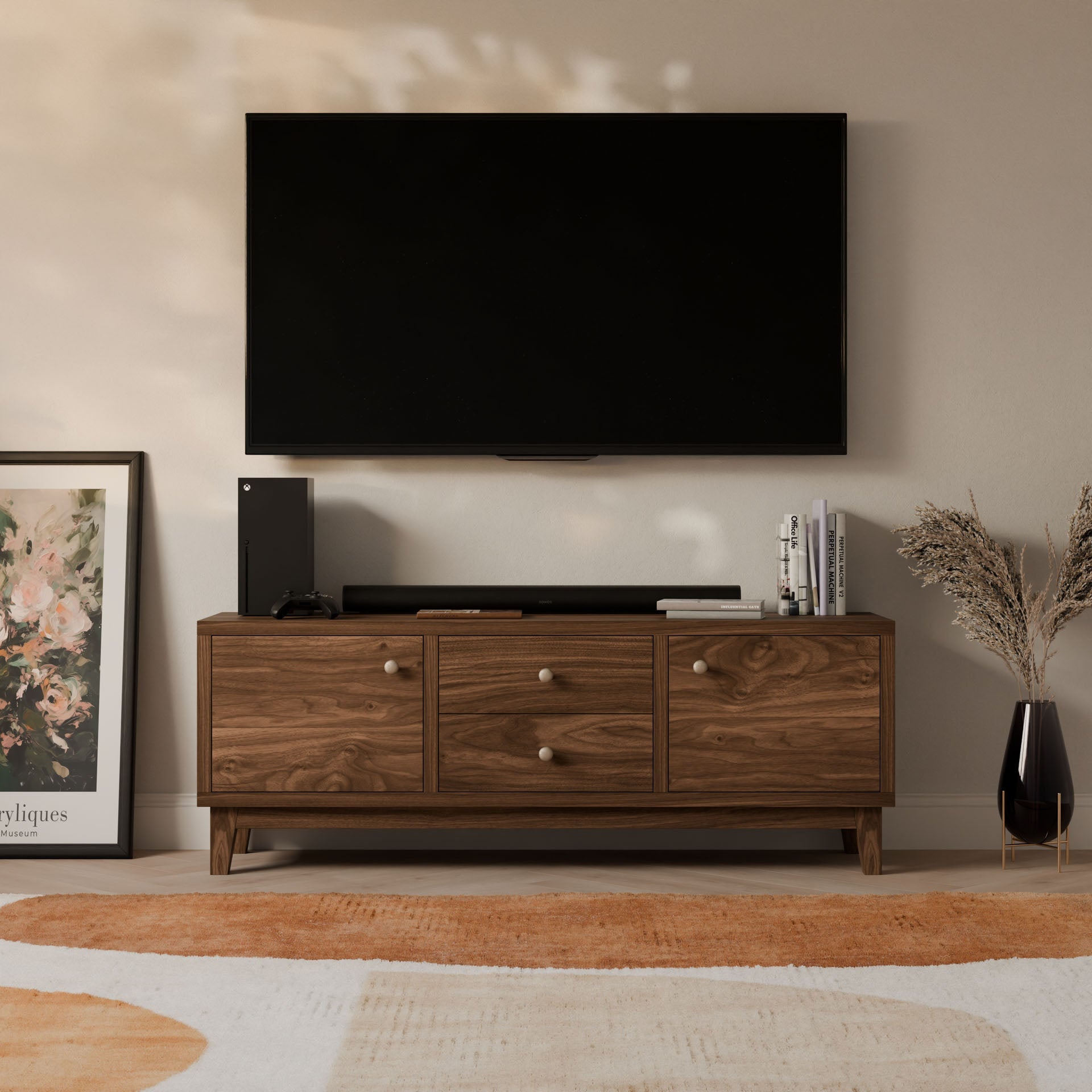 A living room features a wall-mounted TV above a Krovel Furniture Co. Double Drawer, Double Door Media Cabinet in Walnut. The customizable unit holds an Xbox and books, with a stylish framed picture nearby. A vase of dried flowers complements the cozy, patterned rug below.