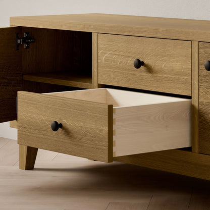 The Double Drawer, Double Door Media Cabinet in White Oak by Krovel Furniture Co. is handcrafted with black knobs, featuring two drawers (one partially open) and an ajar cabinet door, made from eco-friendly, formaldehyde-free hardwood and set on angled legs against a plain wall on a light wood floor.